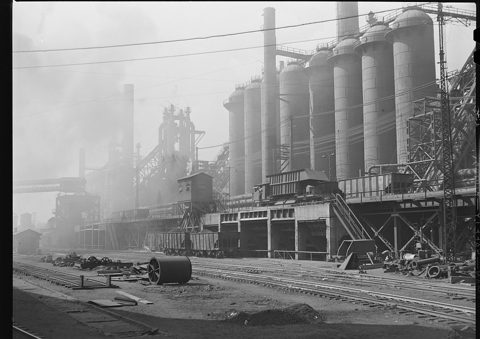 A line of tall cylindrical blast-furnace stoves beside rail tracks, in haze
