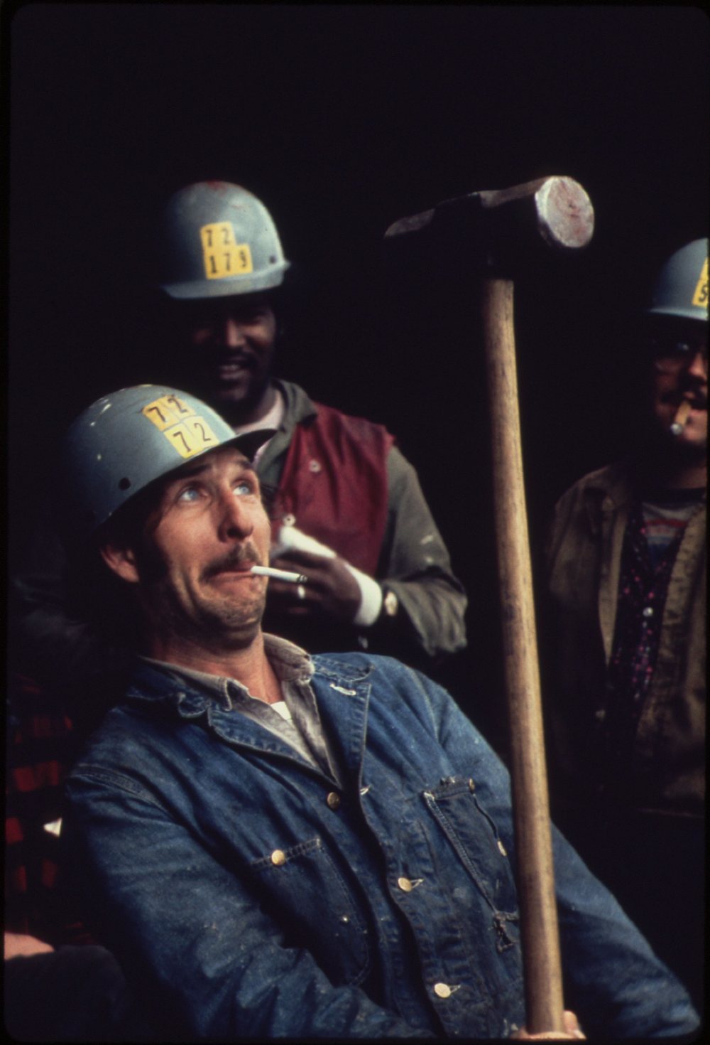 Three shipyard workers in hard hats, one in a denim jacket holding a long-handled sledgehammer and a cigarette