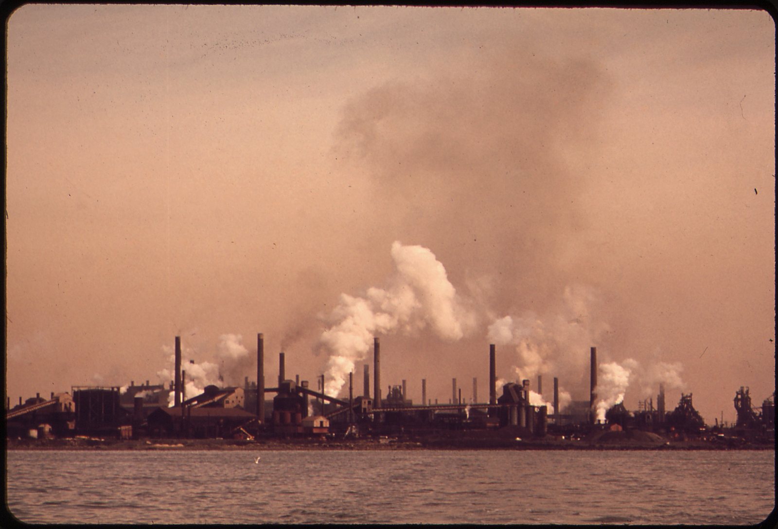 The Bethlehem Steel plant at Sparrows Point seen across the water in 1973: a long line of stacks and plumes of white smoke under a brown sky