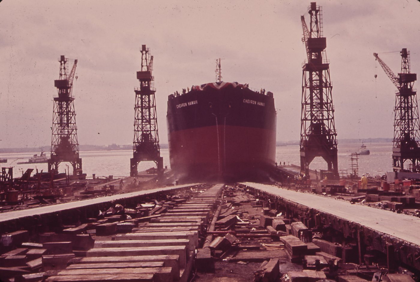 The bow of a large tanker sliding down the launching ways between two tall cranes, timbers and rails in the foreground