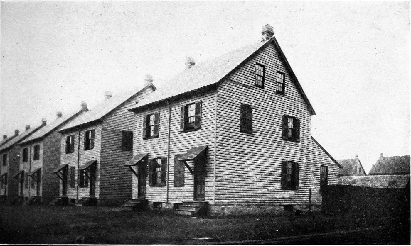 A row of identical two-storey clapboard company houses with steep gables and small porch hoods, on bare ground