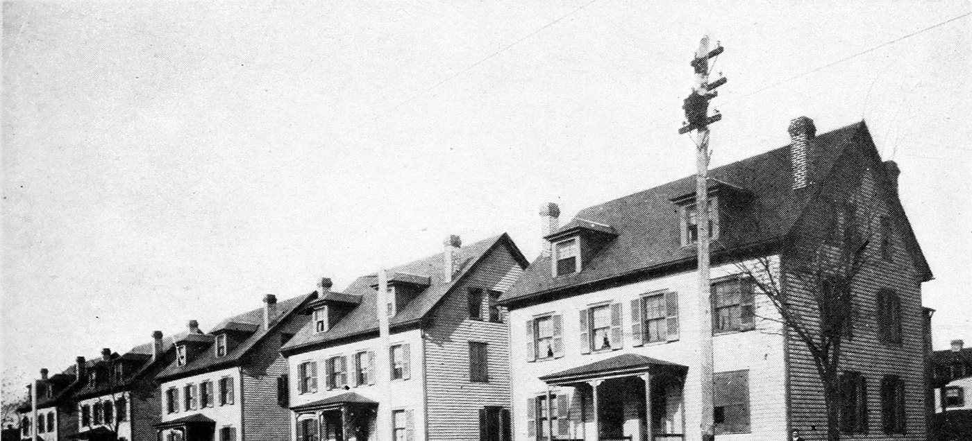 A long terrace of three-storey company houses with dormers and porches along a street, a telegraph pole in front