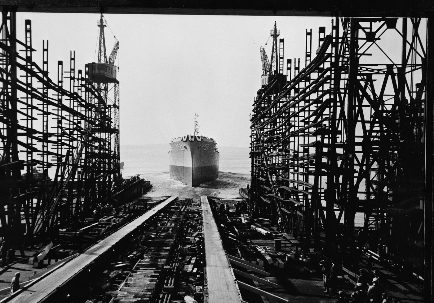 A newly launched ship seen down the empty building ways, between two towers of scaffolding, with spectators below