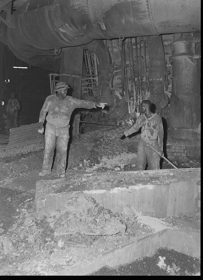 Two men in work clothes standing in a furnace pit under heavy overhead pipes