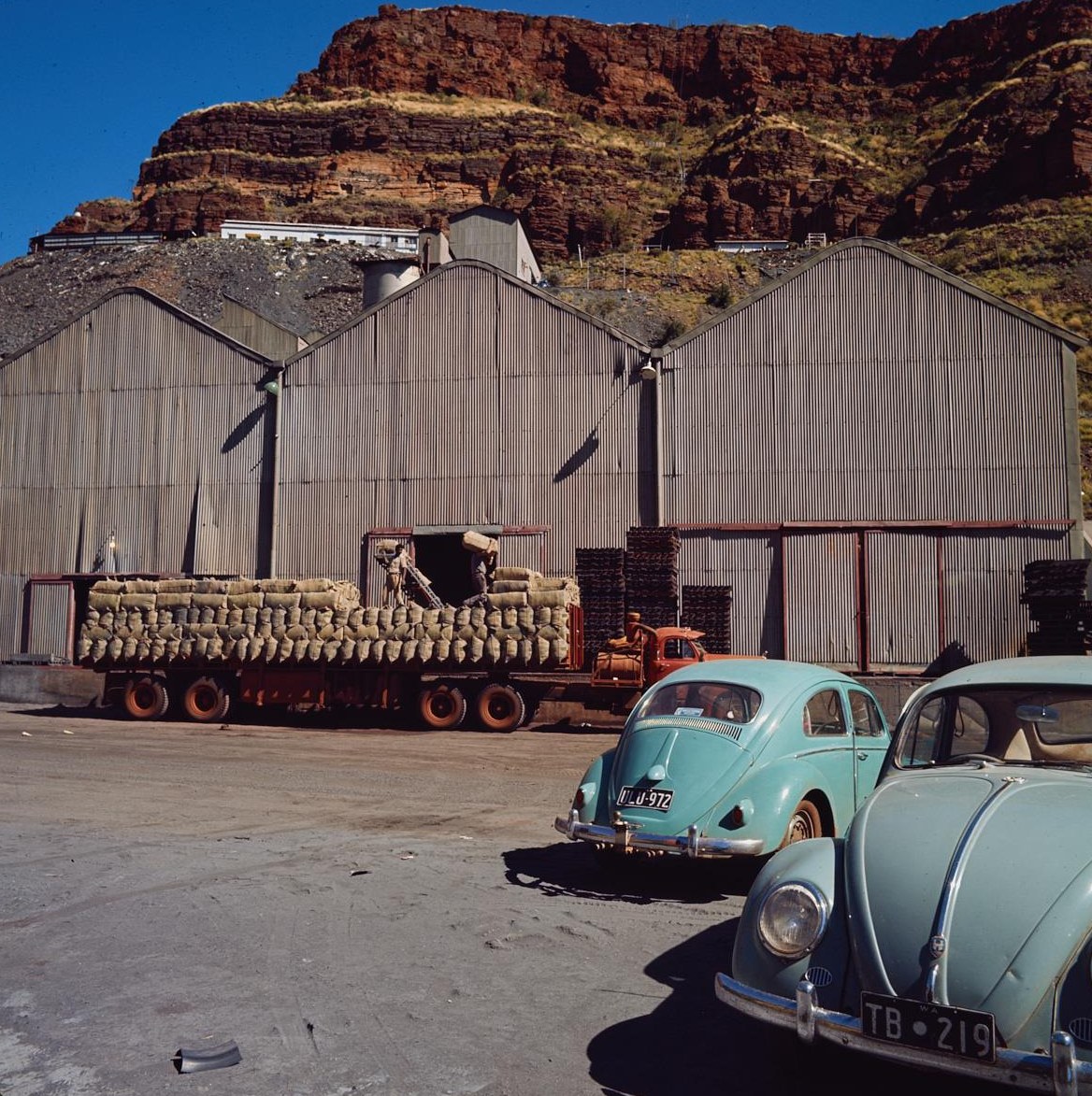 A flatbed truck stacked with sacks of asbestos parked beside large corrugated sheds below red cliffs, two blue Volkswagens in front