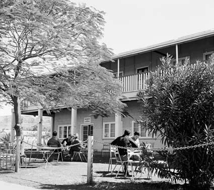 Drinkers at tables in the shade of a tree outside a two-storey hotel with a veranda
