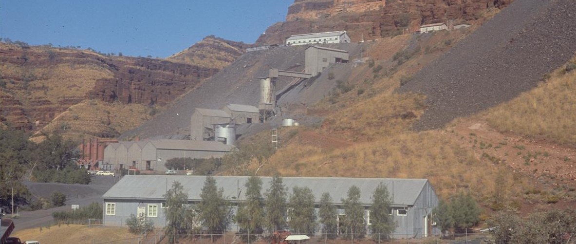 The blue asbestos mill at Wittenoom on a slope of dark tailings below red cliffs, with a long grey shed and a row of trees in front