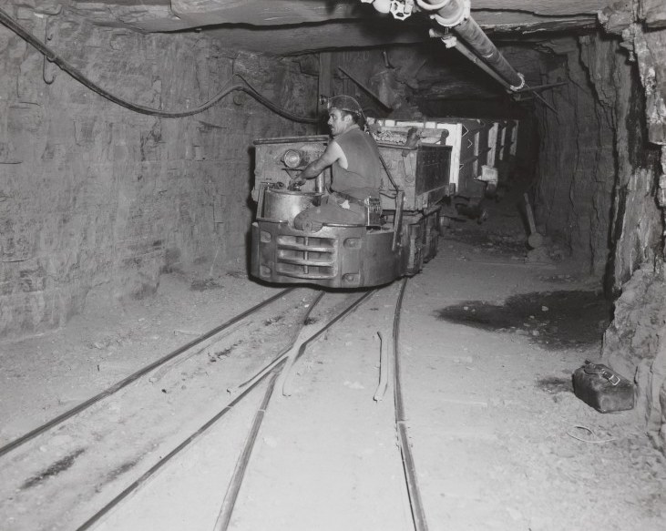 A miner driving a small electric locomotive along narrow rails in a low rock tunnel